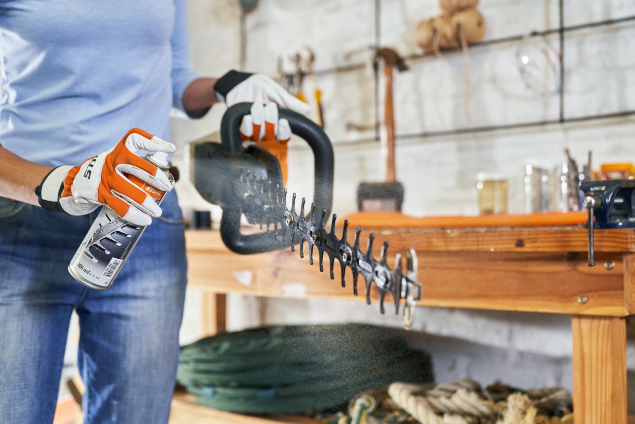 A STIHL HSA 56 cordless hedge trimmer is held by a woman wearing gloves, who is spraying resin solvent onto the cutting apparatus
