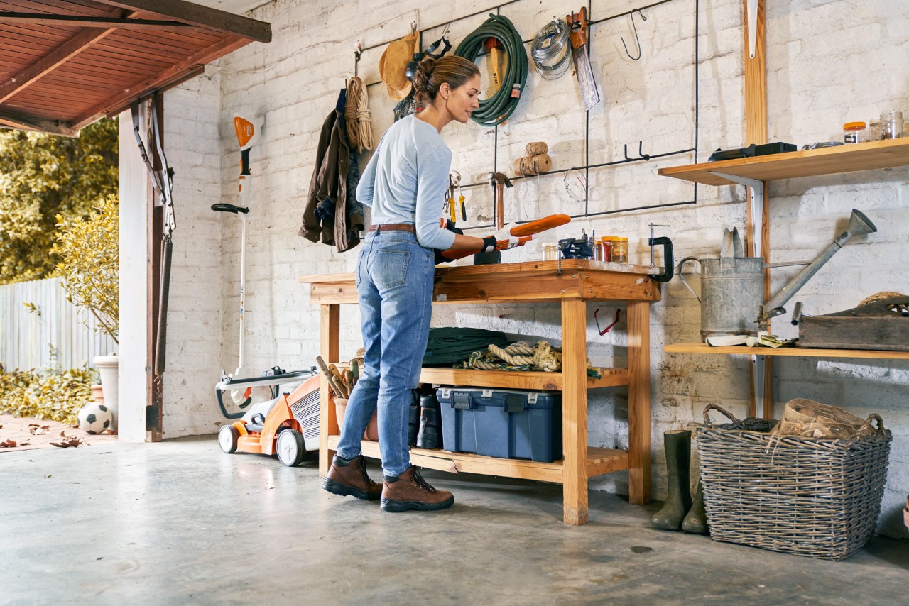 A woman wearing protective gloves stands at a workbench and is working on a STIHL HSA 56 cordless hedge trimmer