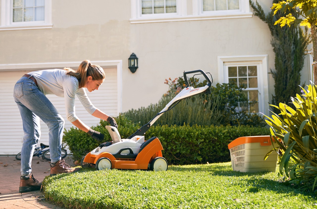 A woman opening the cover of a STIHL RMA 339 C cordless lawn mower in front of a white house