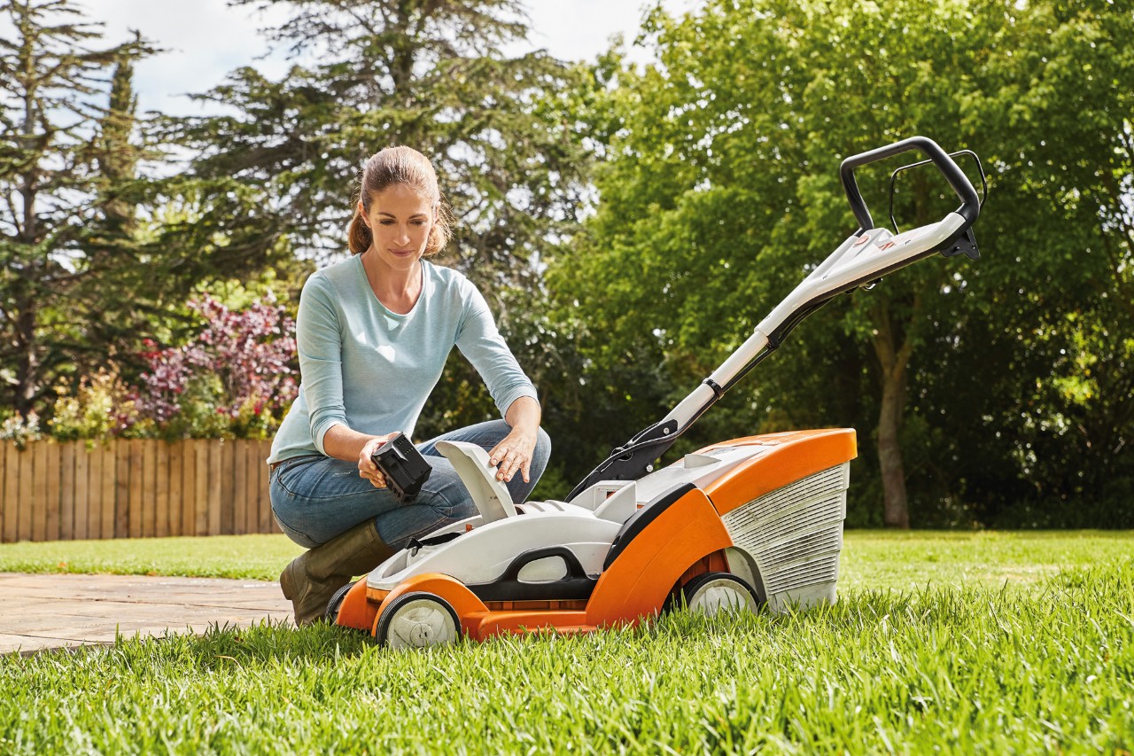 A woman kneeling beside a STIHL RMA 339 C lawn mower and removing the battery from it