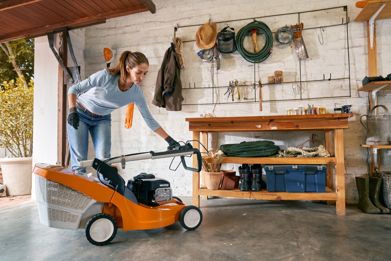 A woman wearing gloves folds down the handlebar of a lawn mower inside a tidy garage 
