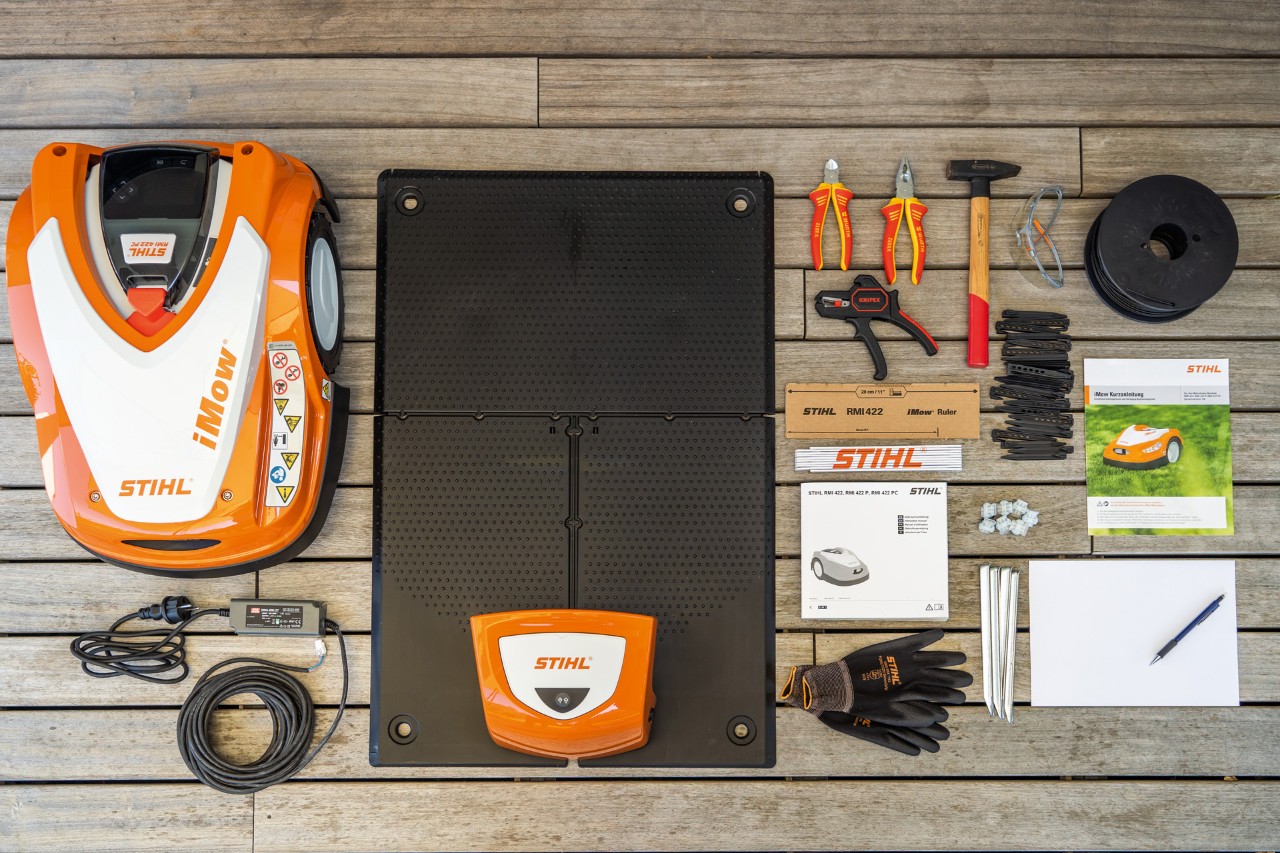 Above shot of an iMOW® robot mower, docking station, cable, gloves, manuals, installation kit and pencil and paper