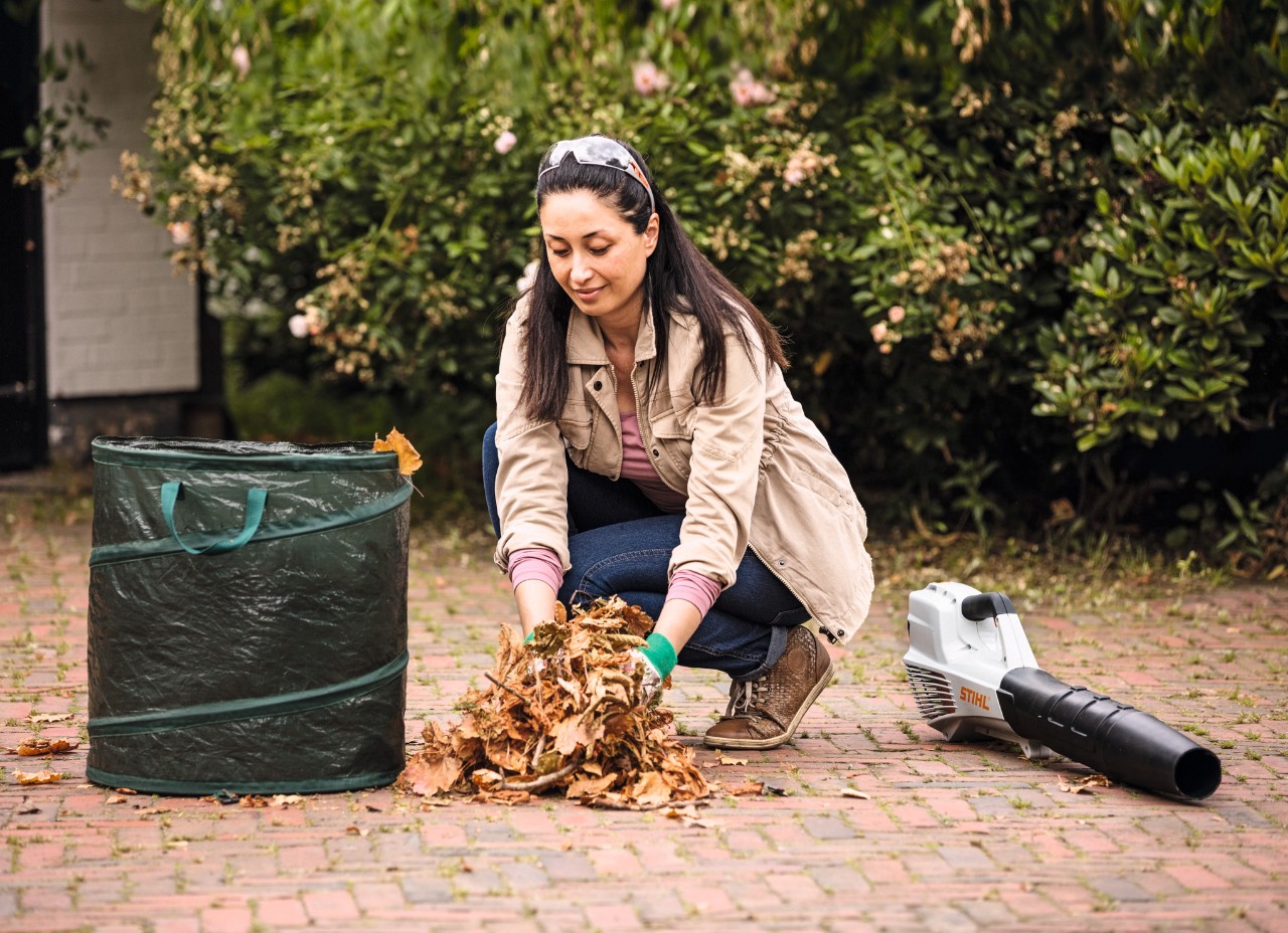 A woman putting leaves into a leaf sack with a STIHL BGA 56 leaf blower on the ground beside her