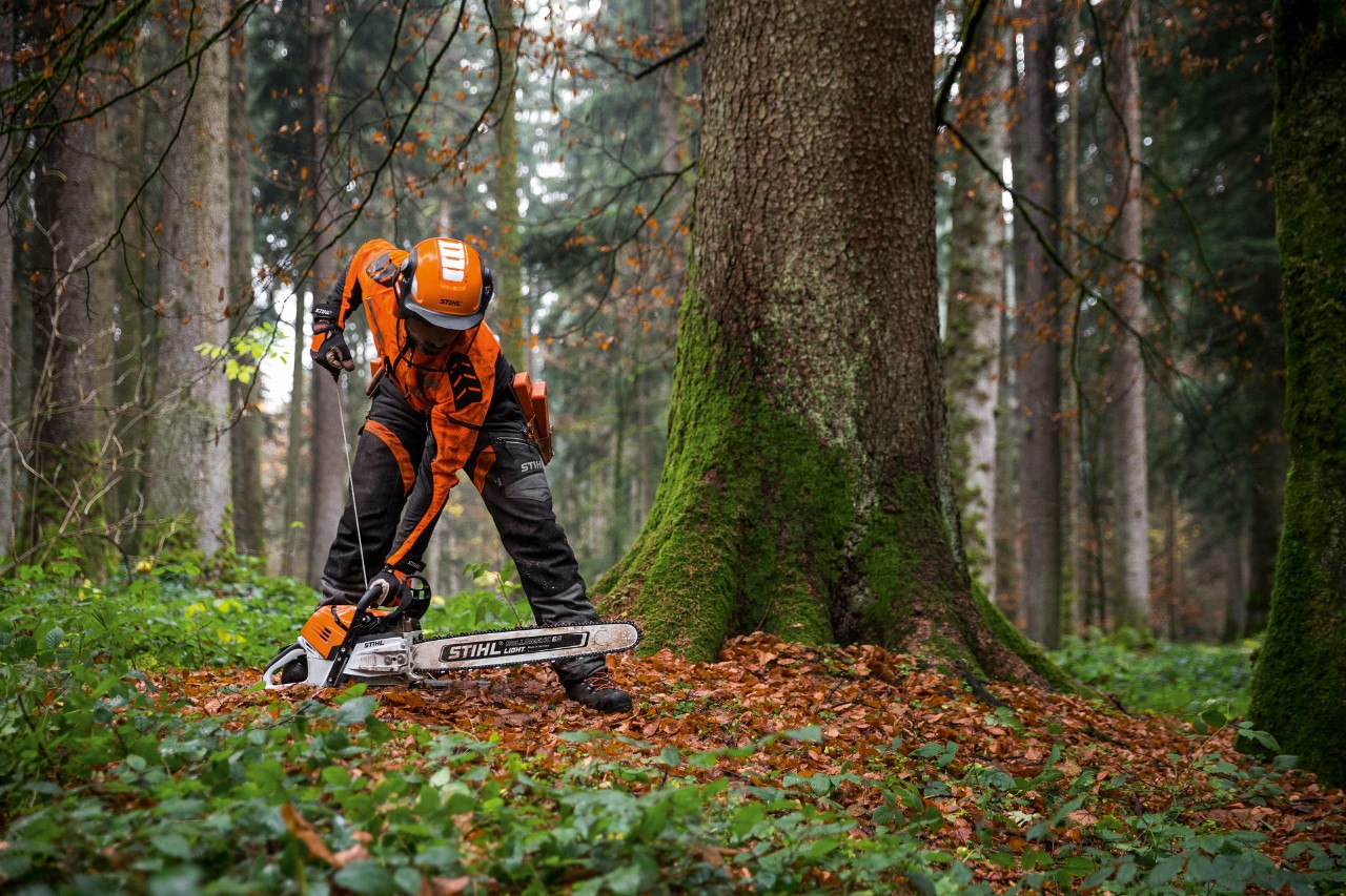 A man in protective clothing starting a STIHL MS 500i petrol chainsaw on the ground, in a deciduous forest.
