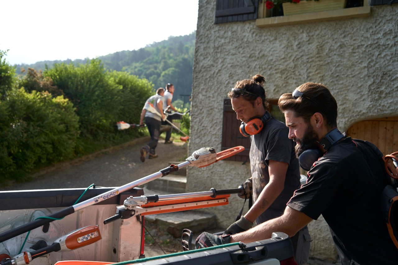 Two landscape gardeners unload STIHL battery power tools from a car, with other workers in the background 