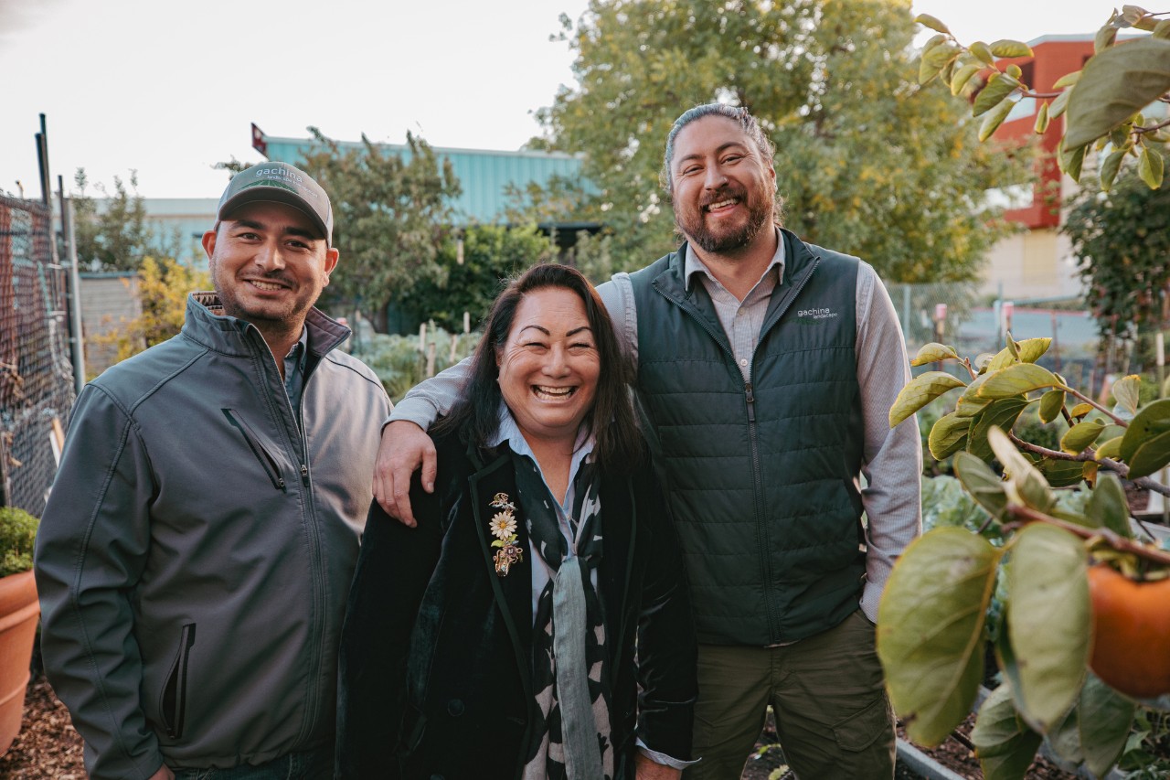 H. Jaclyn Ishimaru-Gachina stands outside in front of her company with her two sons on either side.