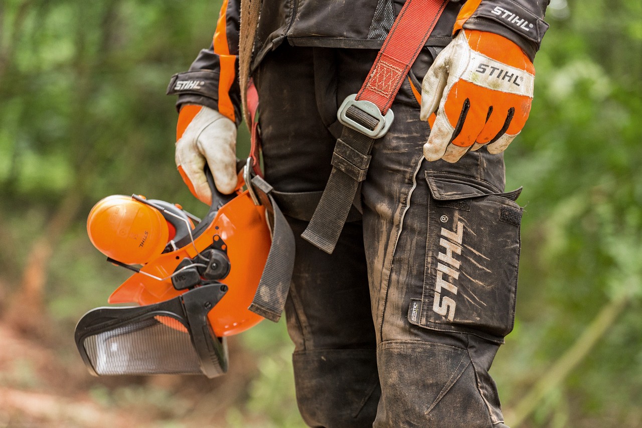 A person wearing STIHL work trousers and gloves, holding a helmet 