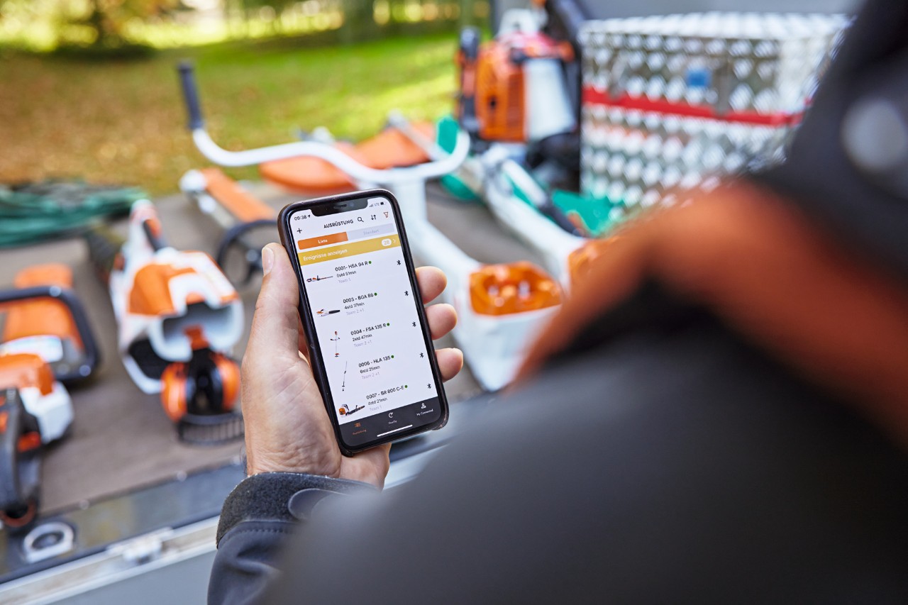 A person standing in front of the loading area of a platform vehicle on which various STIHL tools are laid out. The person holds a smartphone in their hand with the STIHL connected App open. 