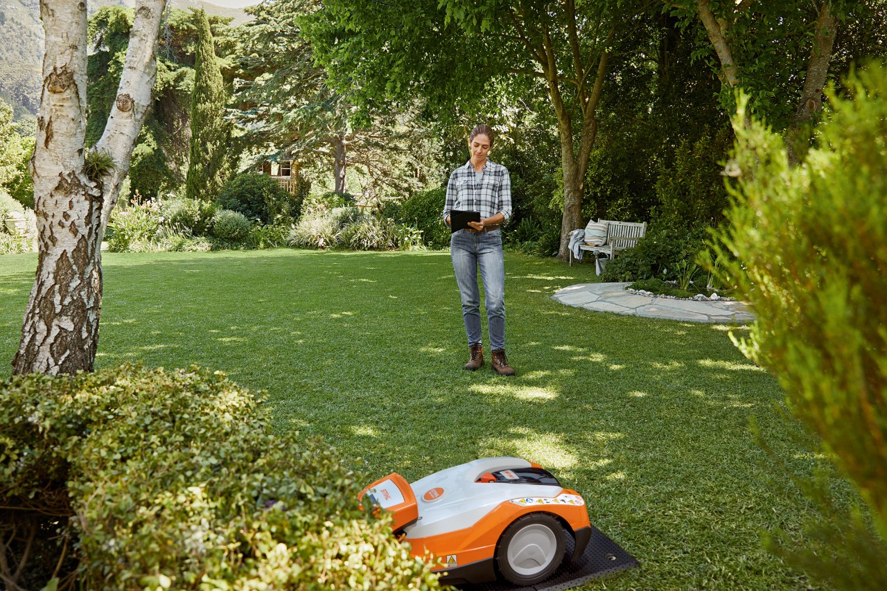 A woman using a tablet in a garden and operating a STIHL iMOW® 522 robot mower