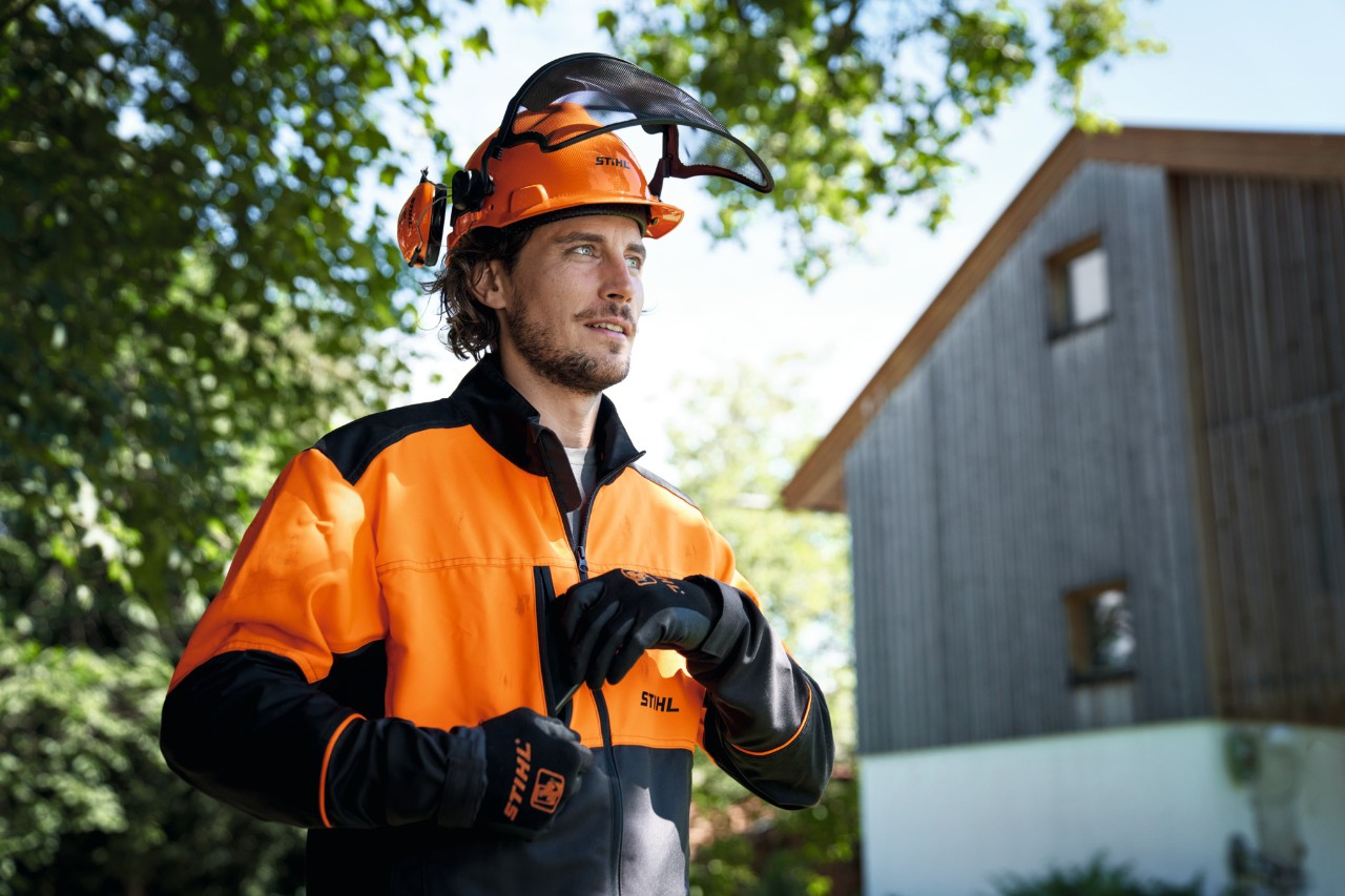 A man wearing orange and black STIHL protective equipment, including a helmet and gloves, looks confidently out of the picture, with a house and trees in the background 