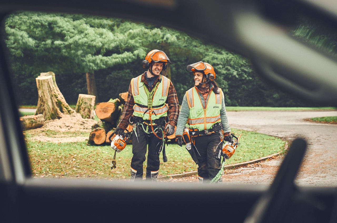 A man and a woman with STIHL cordless chainsaws and a STIHL cordless pruner walk smiling towards a car .